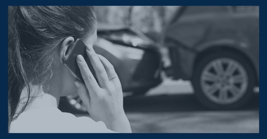 Woman with a cell phone to her ear and a car accident scene in front of her.